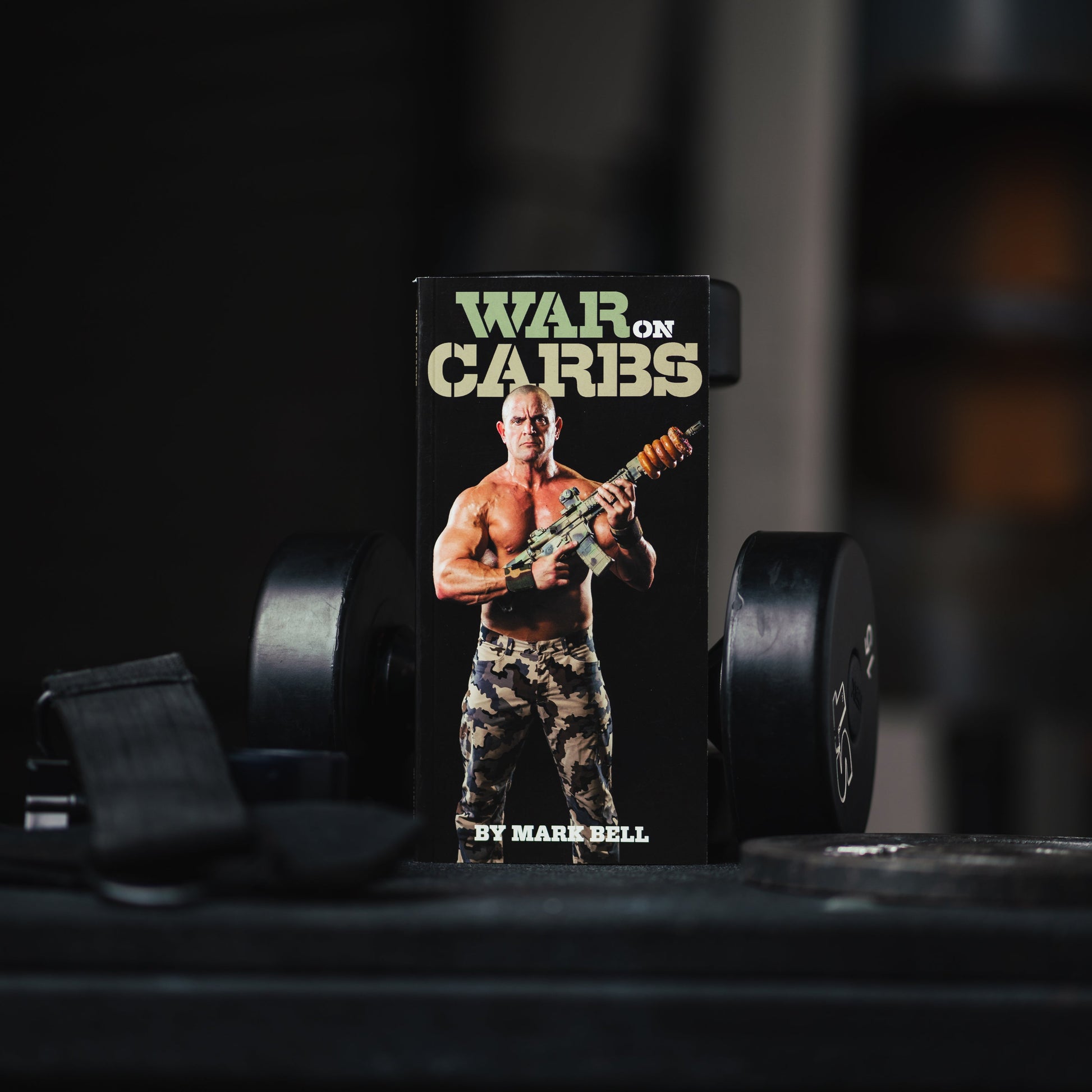 War On Carbs Book by Mark Bell displayed on a gym bench with weights in the background.