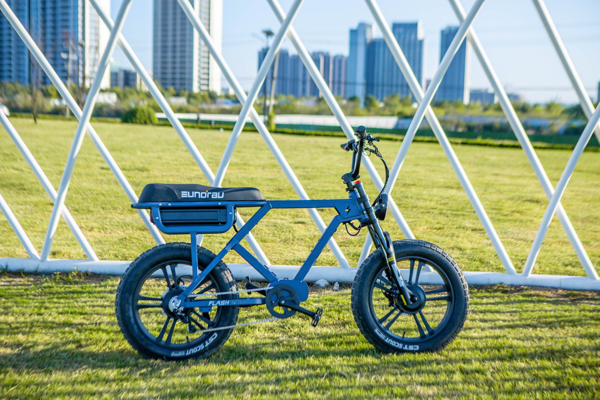 Flash Ebike parked on a grassy area with modern buildings in the background and a geometric structure overhead.