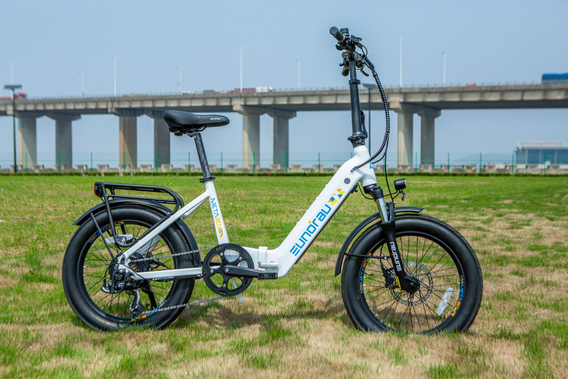 META Foldable City Ebike parked on grass with a bridge in the background, showcasing its compact design and features.