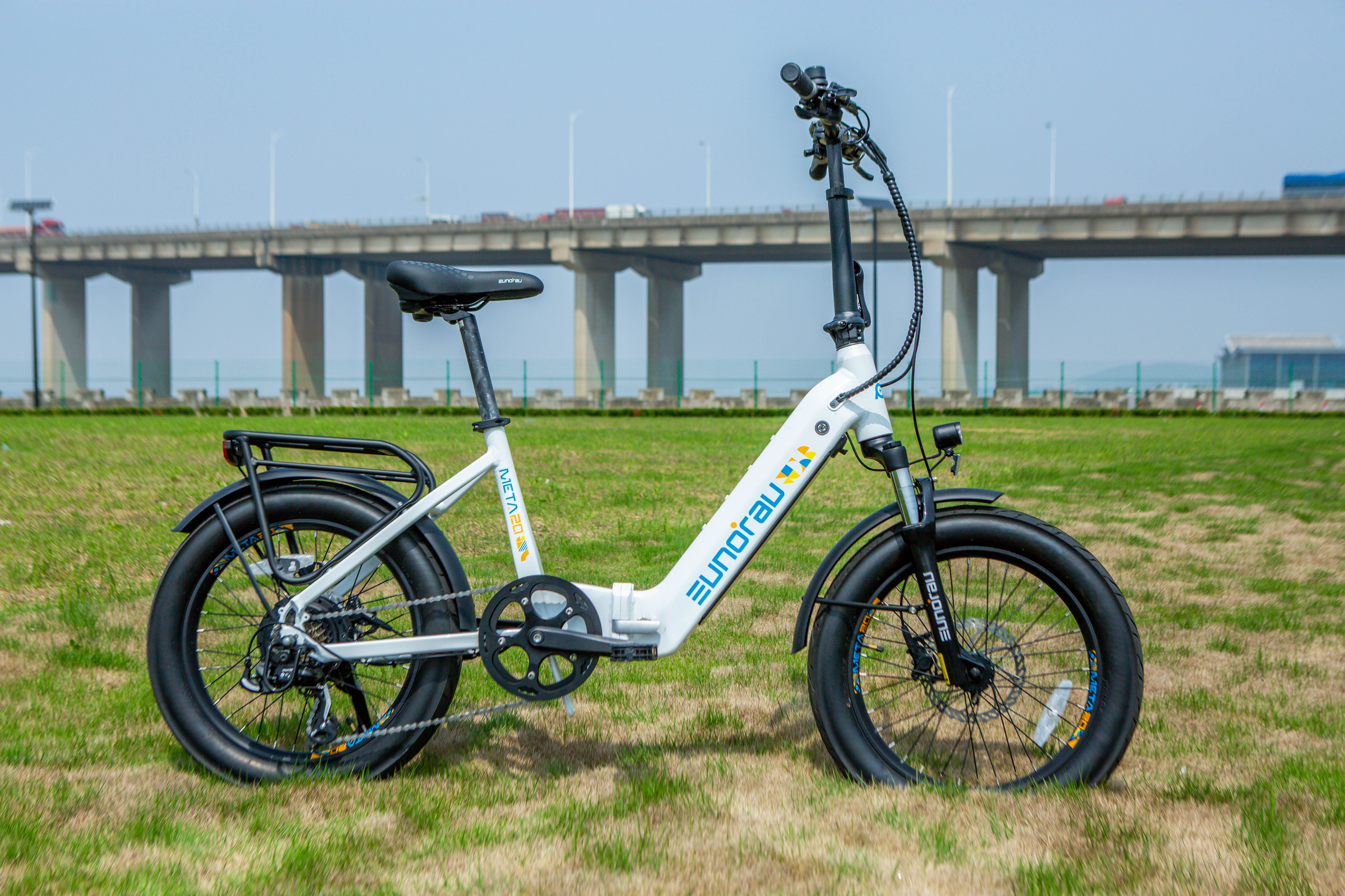 META Foldable City Ebike parked on grass with a bridge in the background, showcasing its compact design and features.